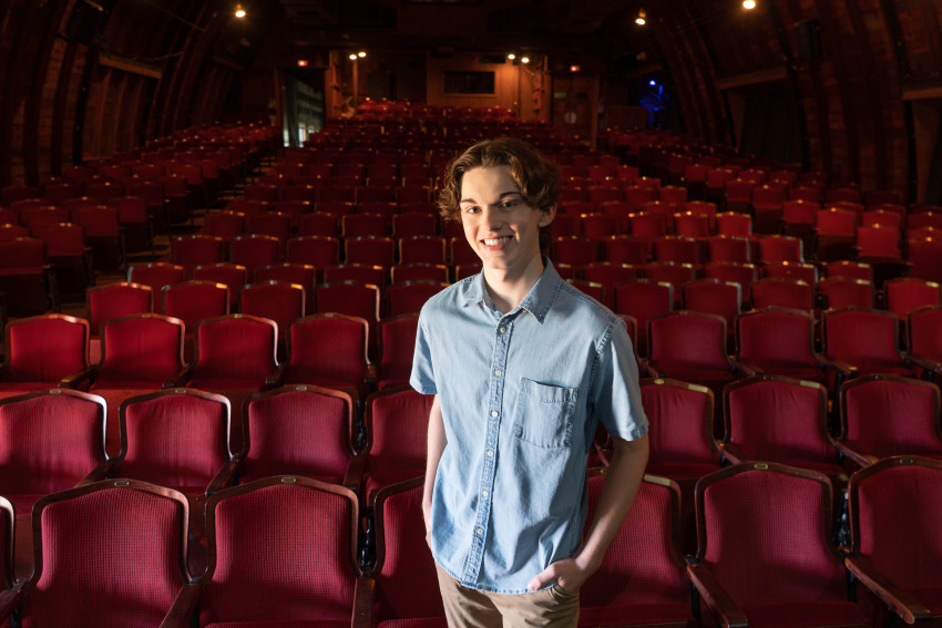 Jack Austin stands in an empty theatre filled with red folding seats.