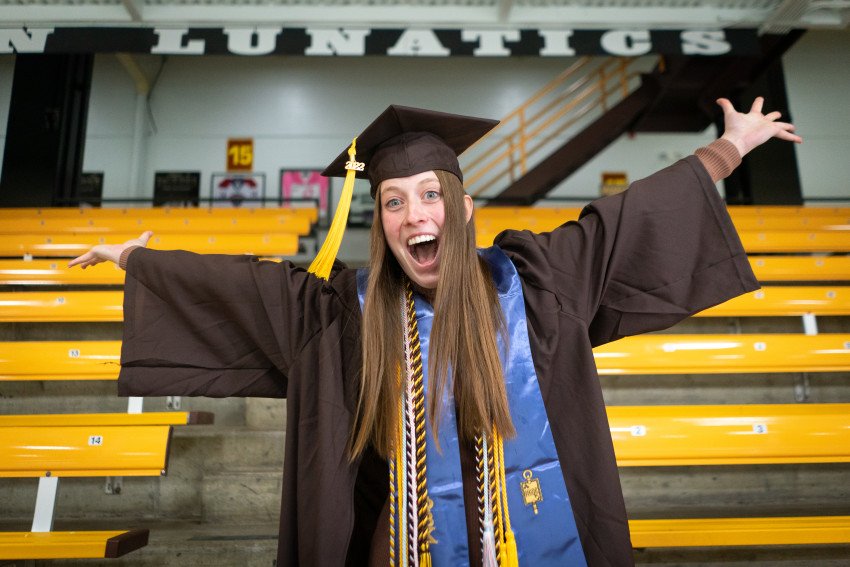 Grace Filpi celebrates with outstretched hands in her cap and gown inside Lawson Arena.