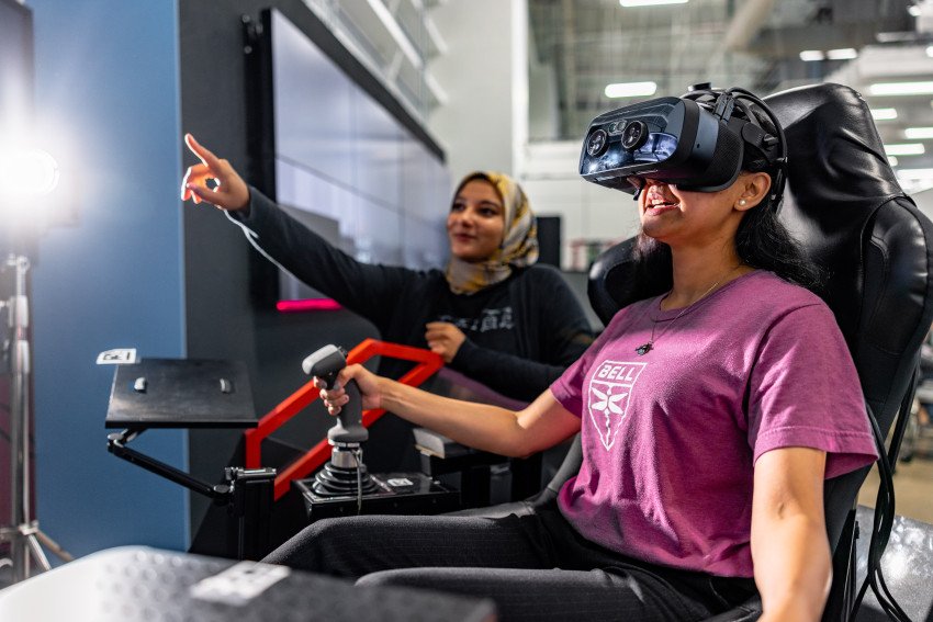 A person wears virtual reality goggles and Aisha Thaj stands behind her pointing at a screen.