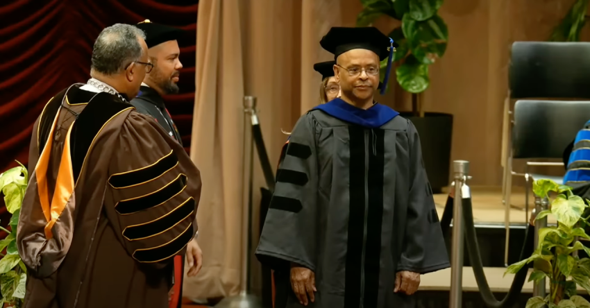 James Rhodes stands on stage in Miller Auditorium wearing full academic regalia next to President Edward Montgomery and Provost Julian Vasquez Heilig.