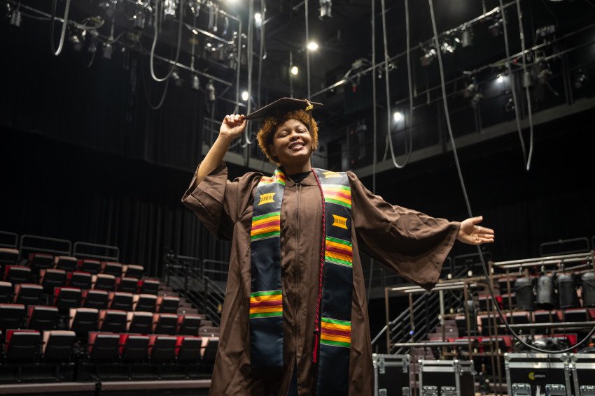Kaide Moore stands in a theatre wearing their graduation regalia.