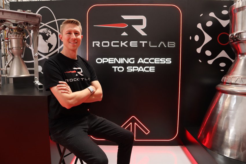 Greg Neff sits with his arms crossed in front of a sign that reads, "Rocket Lab, opening access to space."