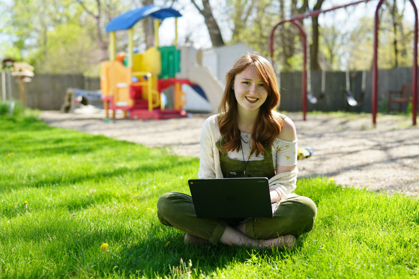 A student sits in the grass holding a laptop in front of a playground for children.