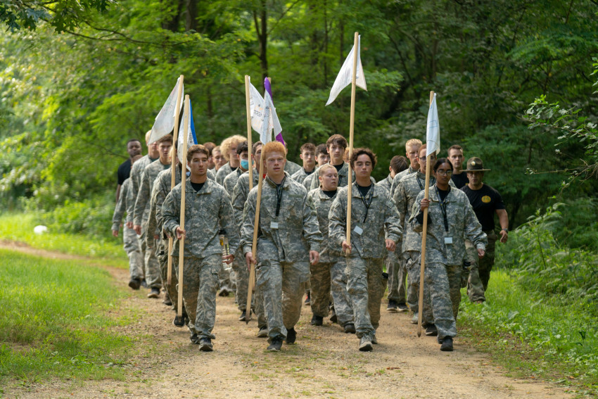 Soldiers walk on a wooded path with flags.