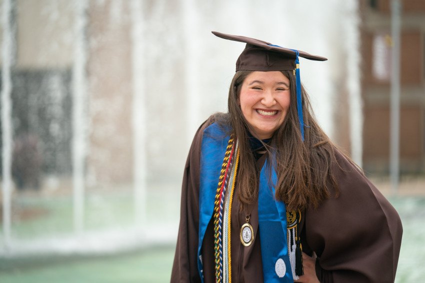 A student smiling in front of Miller Fountain in her graduation cap and gown.