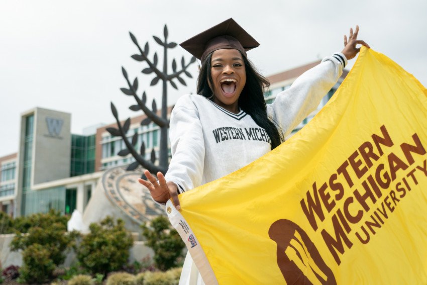 A student wearing a graduation cap holding up a WMU flag in front of the campus seal statue.