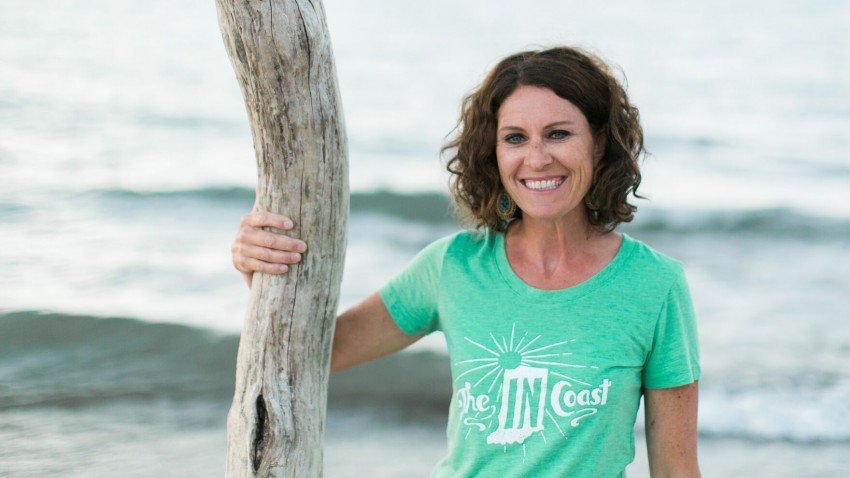 Jennifer Soffin stands on the shoreline wearing a mint green The IN Coast T-shirt.
