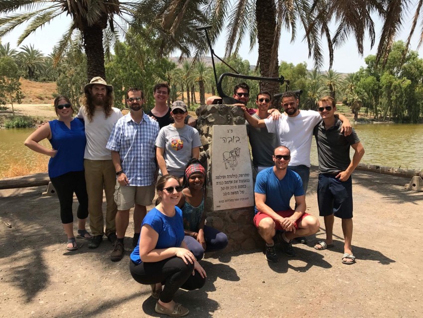 Ten W M U students pose with two other people next to the Jordan river. Three of the students are squatting down in the front while the others stand to each side of a historical marker. The marker is a little larger than an average size person and appears to be made out of stones that have been stacked and fastened together with mortar. A sign is attached to the front of the marker, but it’s slightly out of focus and not in English, so we can’t translate it at this time in this alt tag, but there is a horse depicted on the sign with a harness that looks like it was used for hauling a loaded wagon or perhaps river barge. 
