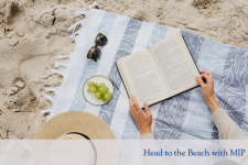 An image of a blanket on sand, with a hat, sunglasses, and two hands holding an open book. 